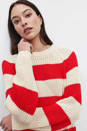 A person wearing the CIARA SWEATER, a red and white striped cotton knit from Velvet by Graham & Spencer, poses with one hand resting on their collar, looking toward the camera.