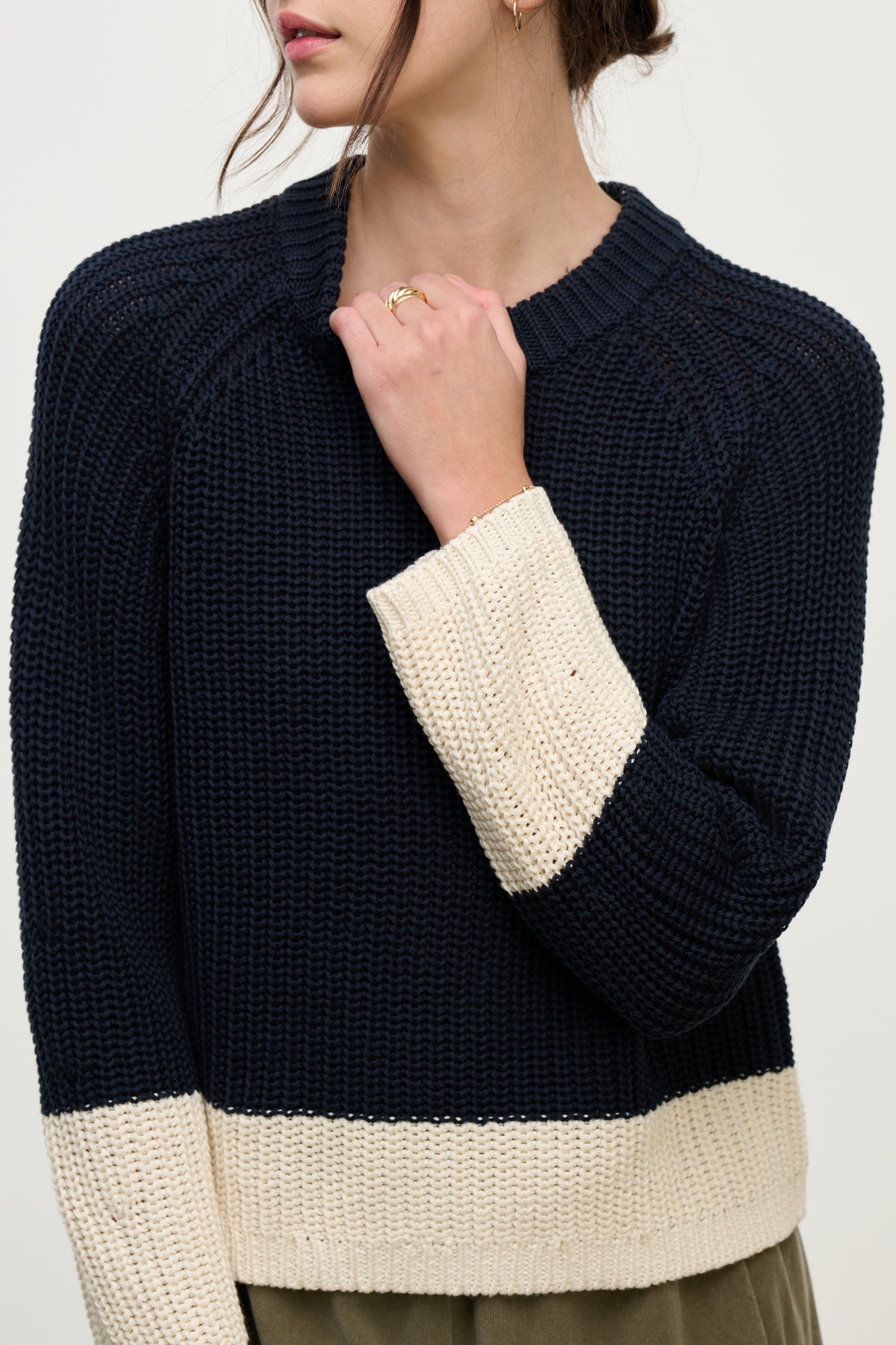 A woman models the SHAY SWEATER by Velvet by Graham & Spencer, featuring a navy blue color block design with cream cuffs and hem, holding her hand near her neck.-39800388354241