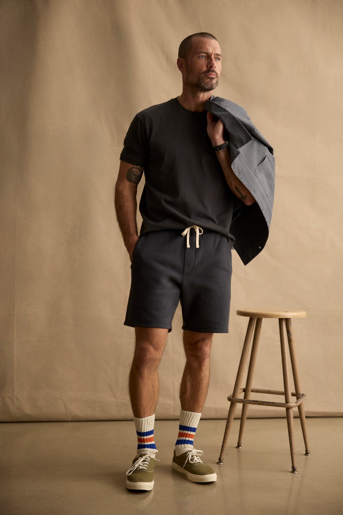 A man stands beside a wooden stool, wearing the ELIAS TEE by Velvet by Graham & Spencer with matching drawstring shorts, striped socks, sneakers, and a gray jacket draped over his shoulder against a plain backdrop.-40606035902657