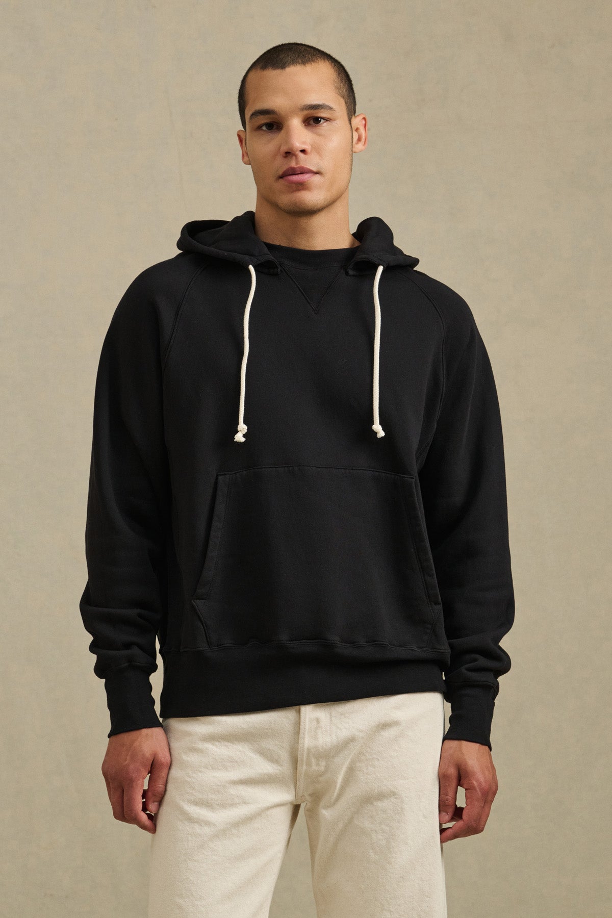 A person faces forward against a neutral background, wearing the BRODIE HOODIE by Velvet by Graham & Spencer—featuring a relaxed fit, drawstring hood, and white accents—paired with off-white pants.-40850658427073