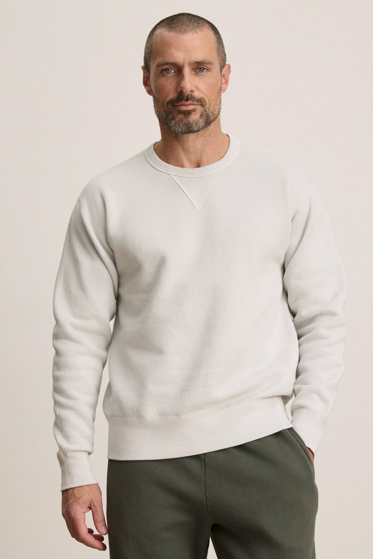 A man with a short beard and buzz cut wears the CILLIAN SWEATSHIRT by Velvet by Graham & Spencer in plain white, paired with olive green pants, standing against a neutral background.