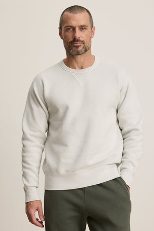 A man with a short beard and buzz cut wears the CILLIAN SWEATSHIRT by Velvet by Graham & Spencer in plain white, paired with olive green pants, standing against a neutral background.