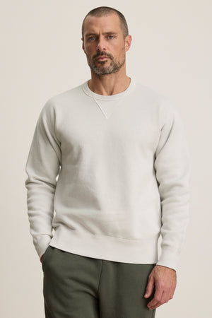 A man with short hair and a beard wears the Velvet by Graham & Spencer CILLIAN SWEATSHIRT in plain white with olive green pants, standing against a neutral background.