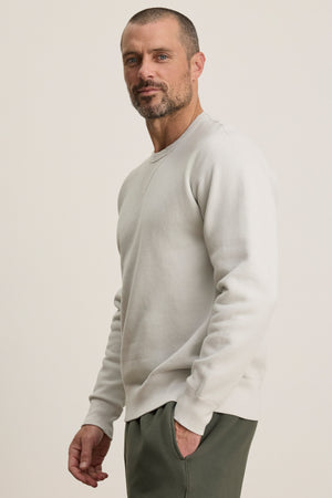A man with short hair and beard wears the CILLIAN SWEATSHIRT by Velvet by Graham & Spencer in light gray, styled with dark green pants, standing against a plain light background.