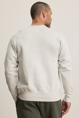 A man with short hair and a neck tattoo faces away, wearing the Velvet by Graham & Spencer CILLIAN SWEATSHIRT—vintage-inspired with a relaxed fit in midweight fleece—paired with dark pants against a plain background.