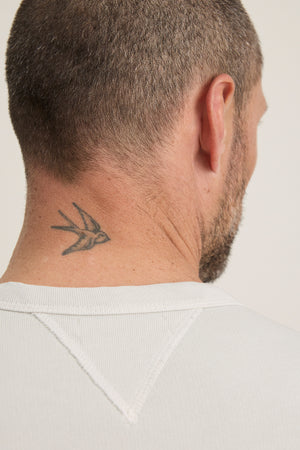 A man with short hair and a beard, seen from behind with a small swallow tattoo on his neck, is wearing the CILLIAN SWEATSHIRT by Velvet by Graham & Spencer—a vintage-inspired, relaxed fit in classic white ribbed style.