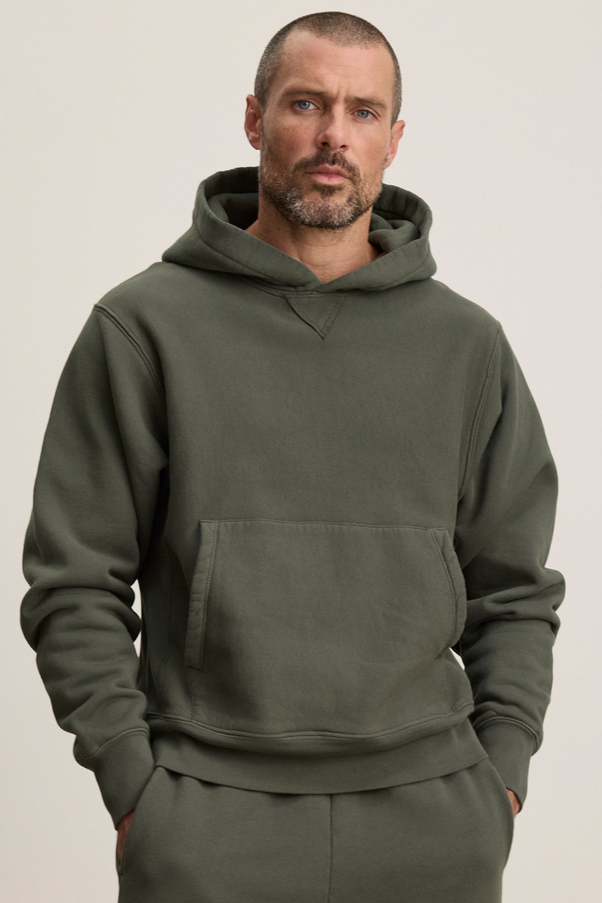 A man with short hair and a beard wears the Velvet by Graham & Spencer KAZ HOODIE, a dark green midweight fleece hoodie with a relaxed fit, kangaroo pocket, and matching pants, standing against a plain light background.
