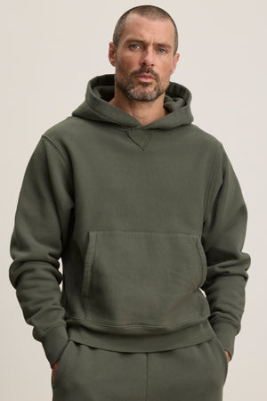 A man with short hair and a beard wears the Velvet by Graham & Spencer KAZ HOODIE, a dark green midweight fleece hoodie with a relaxed fit, kangaroo pocket, and matching pants, standing against a plain light background.