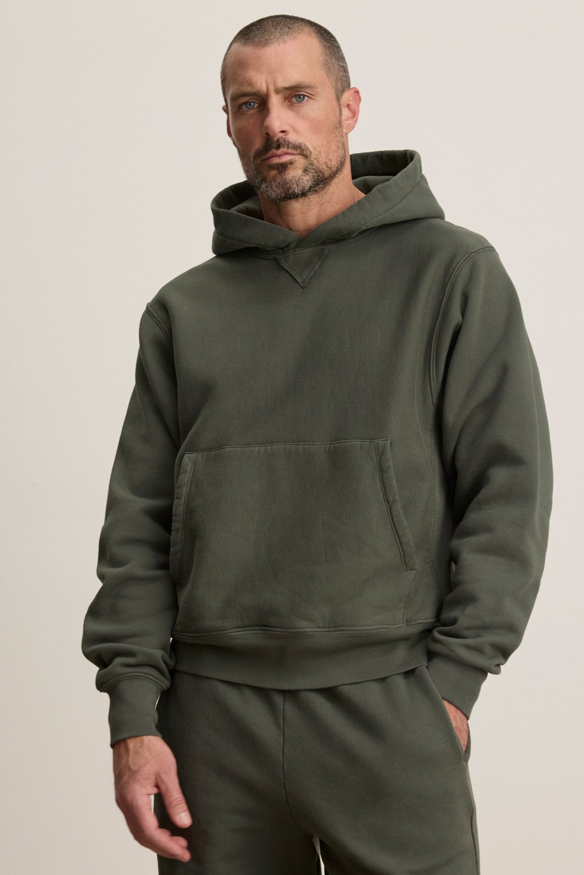 Model wears the KAZ HOODIE by Velvet by Graham & Spencer, an olive green relaxed fit with matching sweatpants and a kangaroo pouch pocket, set against a plain light background.