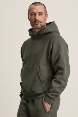 A man with short hair and a beard wears the KAZ HOODIE by Velvet by Graham & Spencer with matching pants, standing with one hand in his pocket against a plain background.