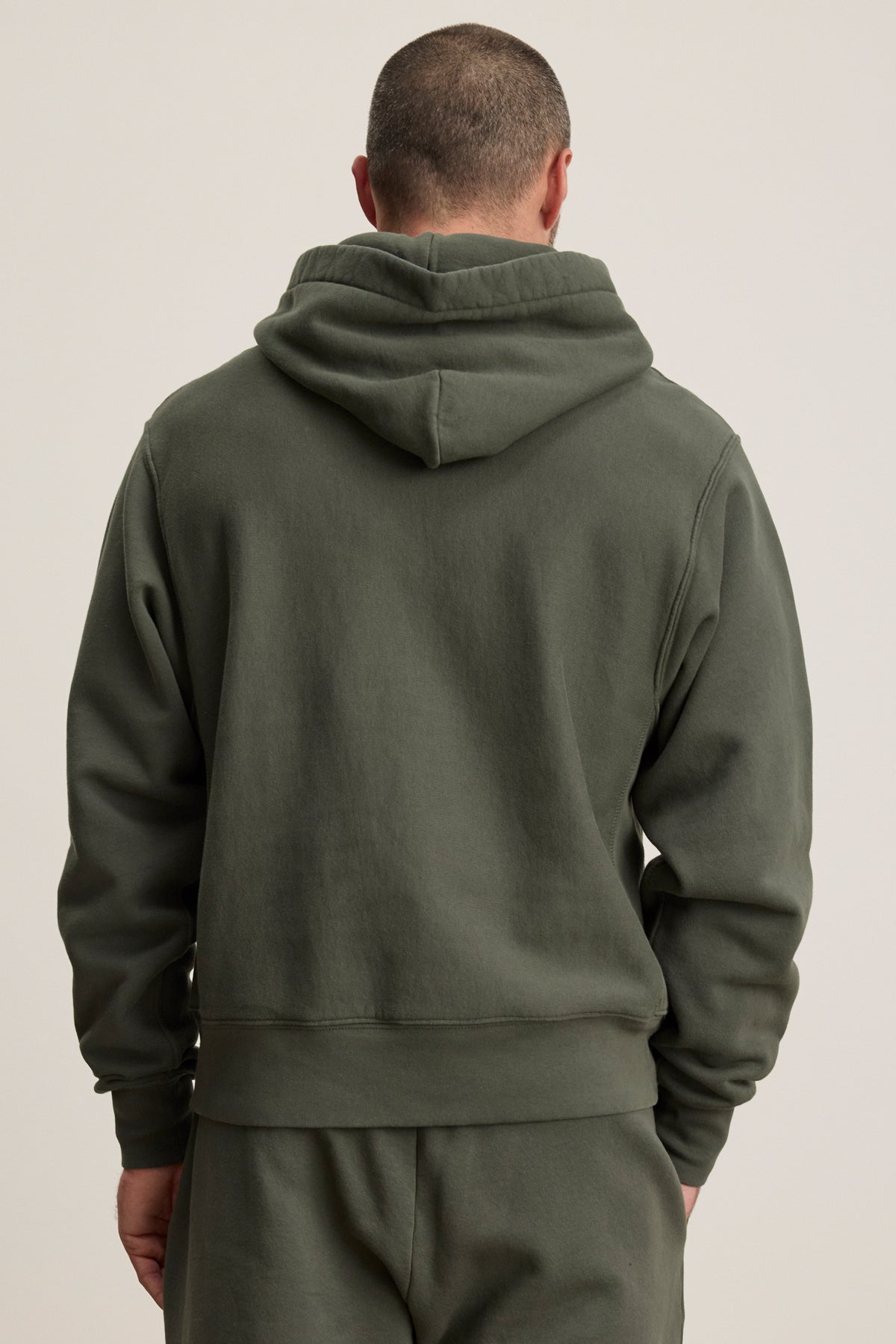 A person is seen from the back wearing the Velvet by Graham & Spencer KAZ HOODIE in olive green with a relaxed fit, kangaroo pouch pocket, and matching pants, set against a plain background.