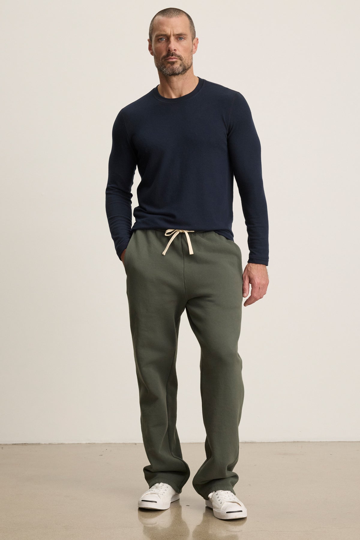 A man stands against a plain background wearing a navy long-sleeve shirt, Velvet by Graham & Spencer HUDSON SWEATPANT in olive green cotton fleece, and white sneakers—ideal for everyday comfort.