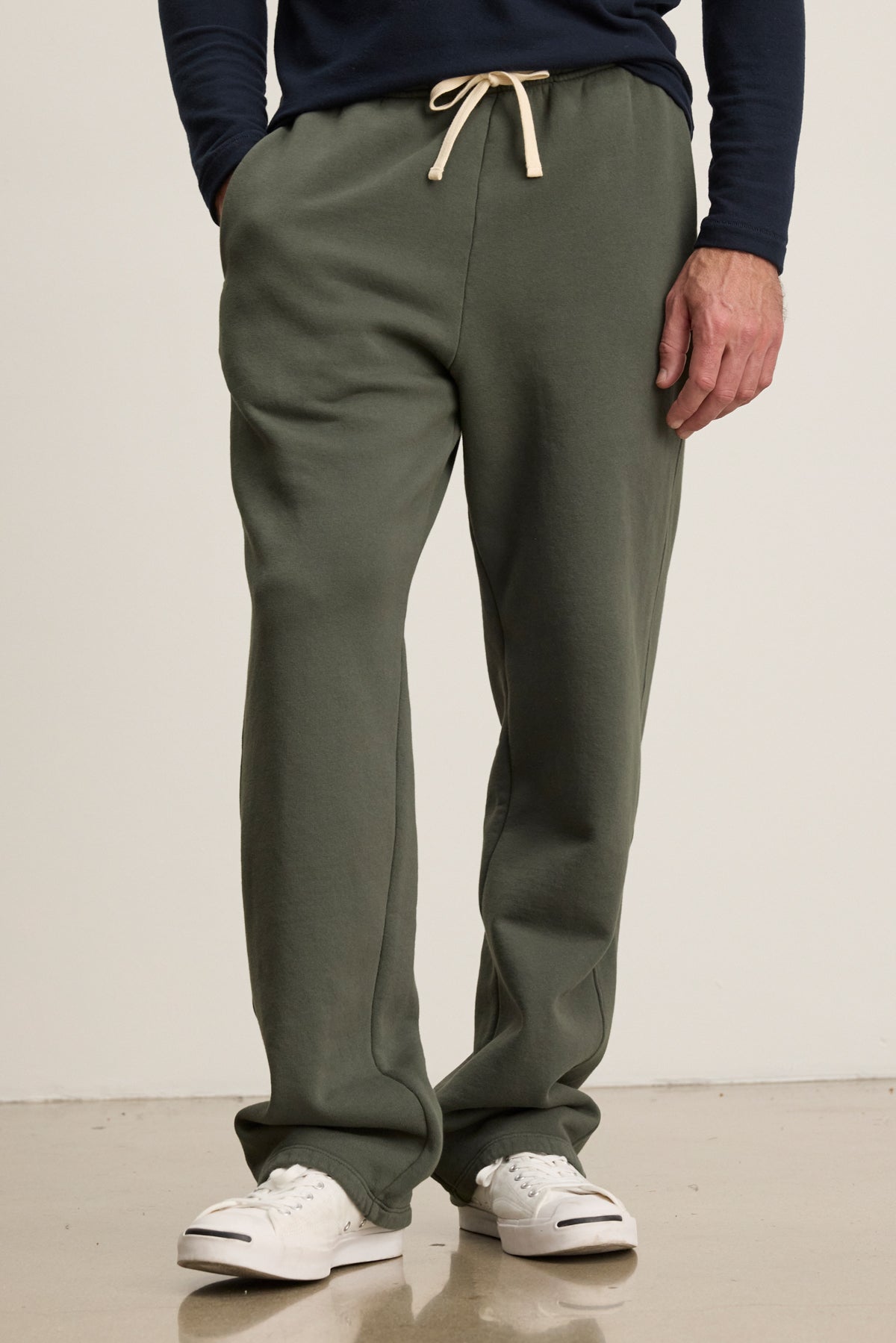Someone stands on a light floor wearing Velvet by Graham & Spencer’s HUDSON SWEATPANT in olive green cotton fleece with a beige drawstring, paired with a navy long-sleeve shirt and white sneakers—ideal for everyday style.