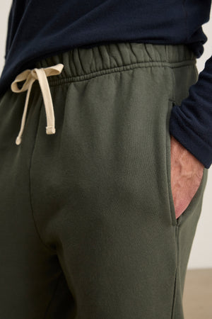 A person wears Velvet by Graham & Spencer's HUDSON SWEATPANT in dark green cotton fleece and a navy blue long-sleeve shirt, one hand in their pocket—an ideal look for everyday wear.