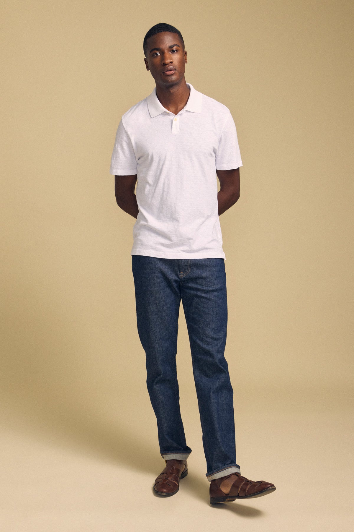 A man stands against a beige background, wearing the MORRIS POLO by Velvet by Graham & Spencer—a white cotton slub polo with short sleeves and a ribbed collar—paired with dark blue jeans and brown shoes, his hands behind his back.-41702098239681