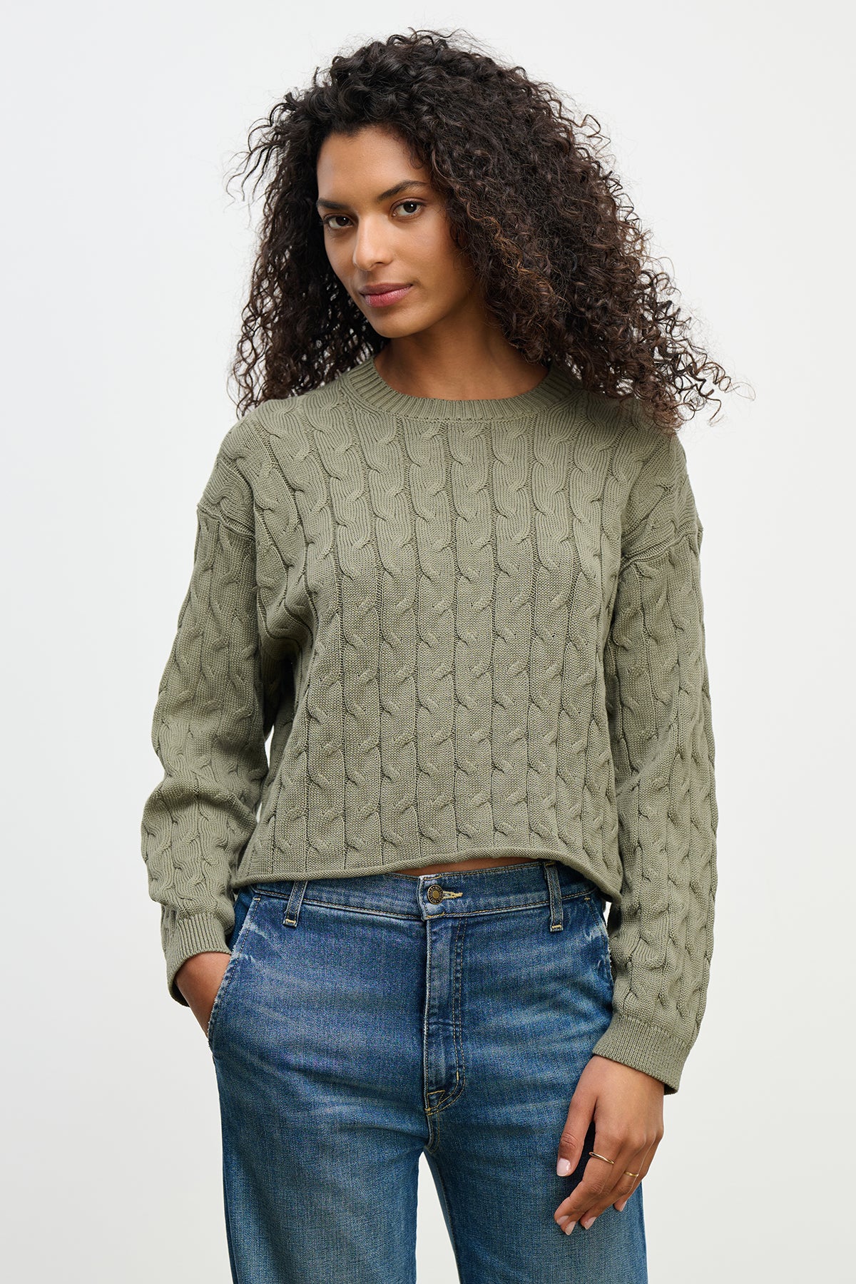 Someone with curly hair wearing the APRIL SWEATER by Velvet by Graham & Spencer, a green cable-knit cotton sweater in a boxy fit, and blue jeans, stands against a plain background.