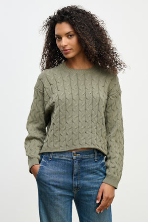 Someone with curly hair wearing the APRIL SWEATER by Velvet by Graham & Spencer, a green cable-knit cotton sweater in a boxy fit, and blue jeans, stands against a plain background.