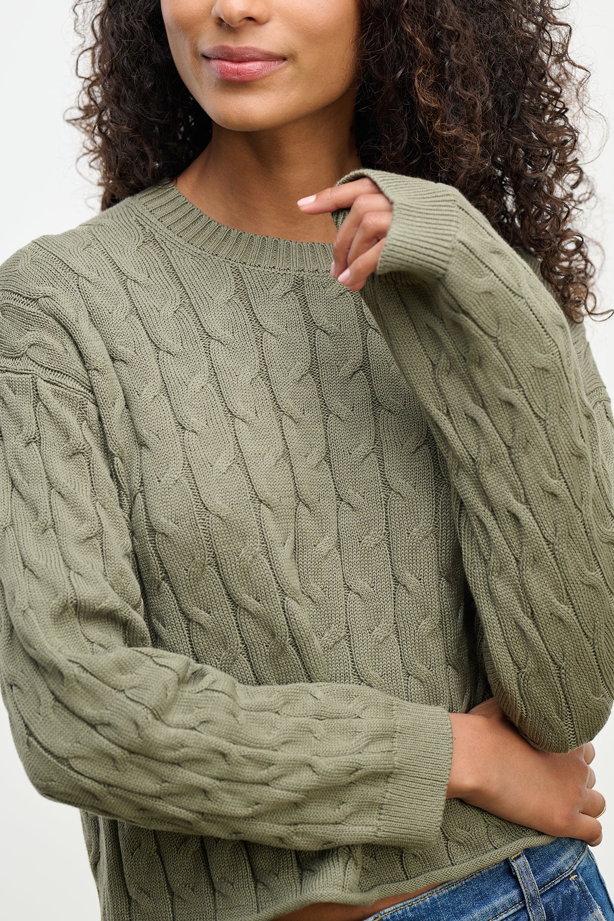 A person wearing the APRIL SWEATER by Velvet by Graham & Spencer, featuring a boxy-fit green cable-knit cotton design and long sleeves, crossing their arms. Only their upper body is visible.