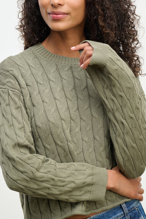 A person wearing the APRIL SWEATER by Velvet by Graham & Spencer, featuring a boxy-fit green cable-knit cotton design and long sleeves, crossing their arms. Only their upper body is visible.