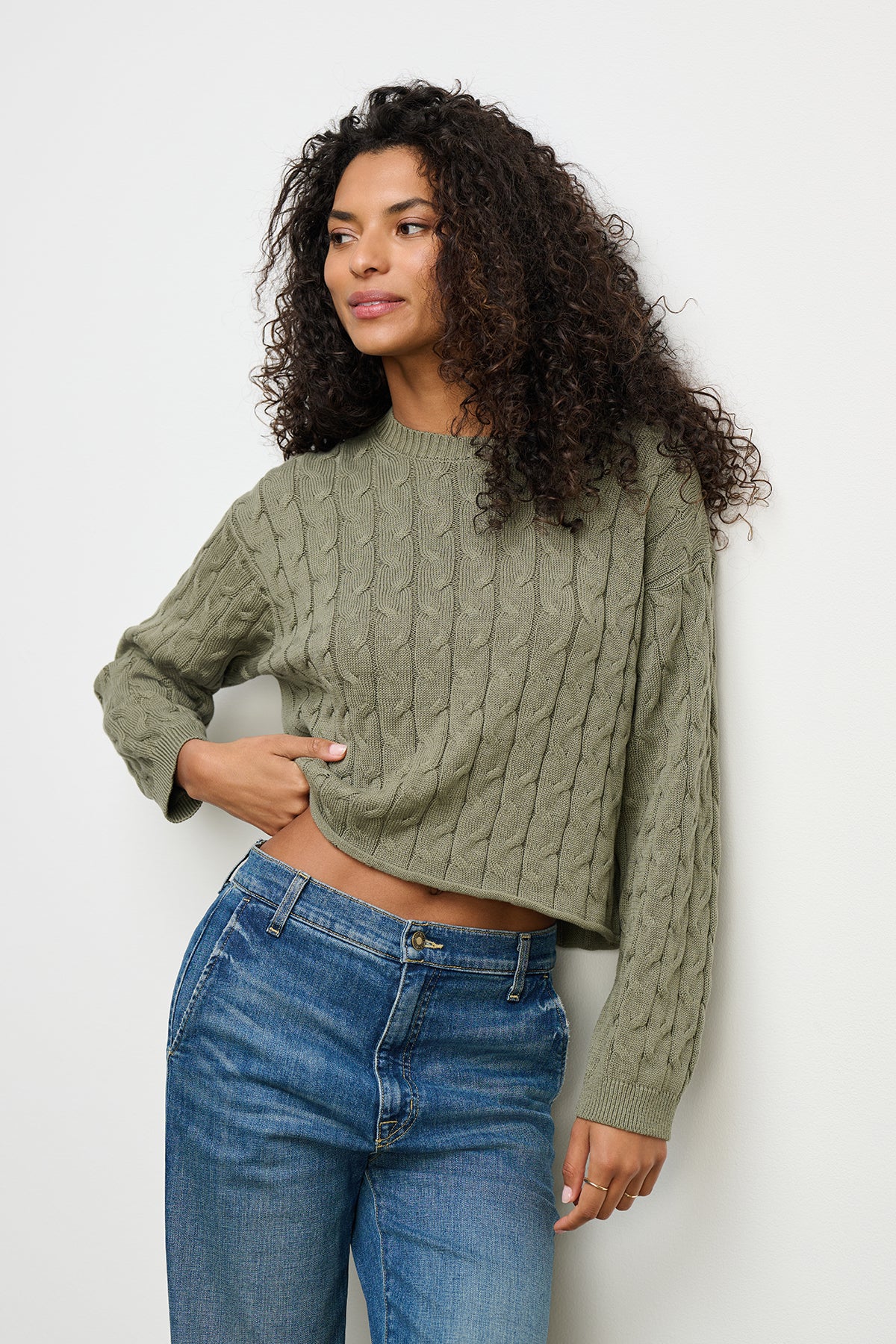 A person with curly hair wears the APRIL SWEATER by Velvet by Graham & Spencer, a green boxy-fit cable-knit style, paired with blue jeans, standing against a light-colored wall.