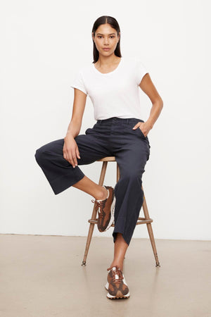 A woman seated on a wooden stool, wearing a white t-shirt and navy garment dye culottes with brown strappy sandals.(Product Name: Velvet by Graham & Spencer MYA COTTON CANVAS PANT.)