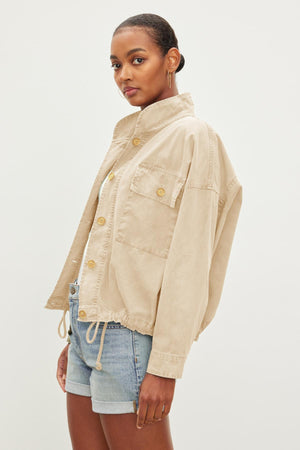 A person wearing a beige NICOLA JACKET by Velvet by Graham & Spencer with buttons and blue denim shorts, posing against a plain white background.