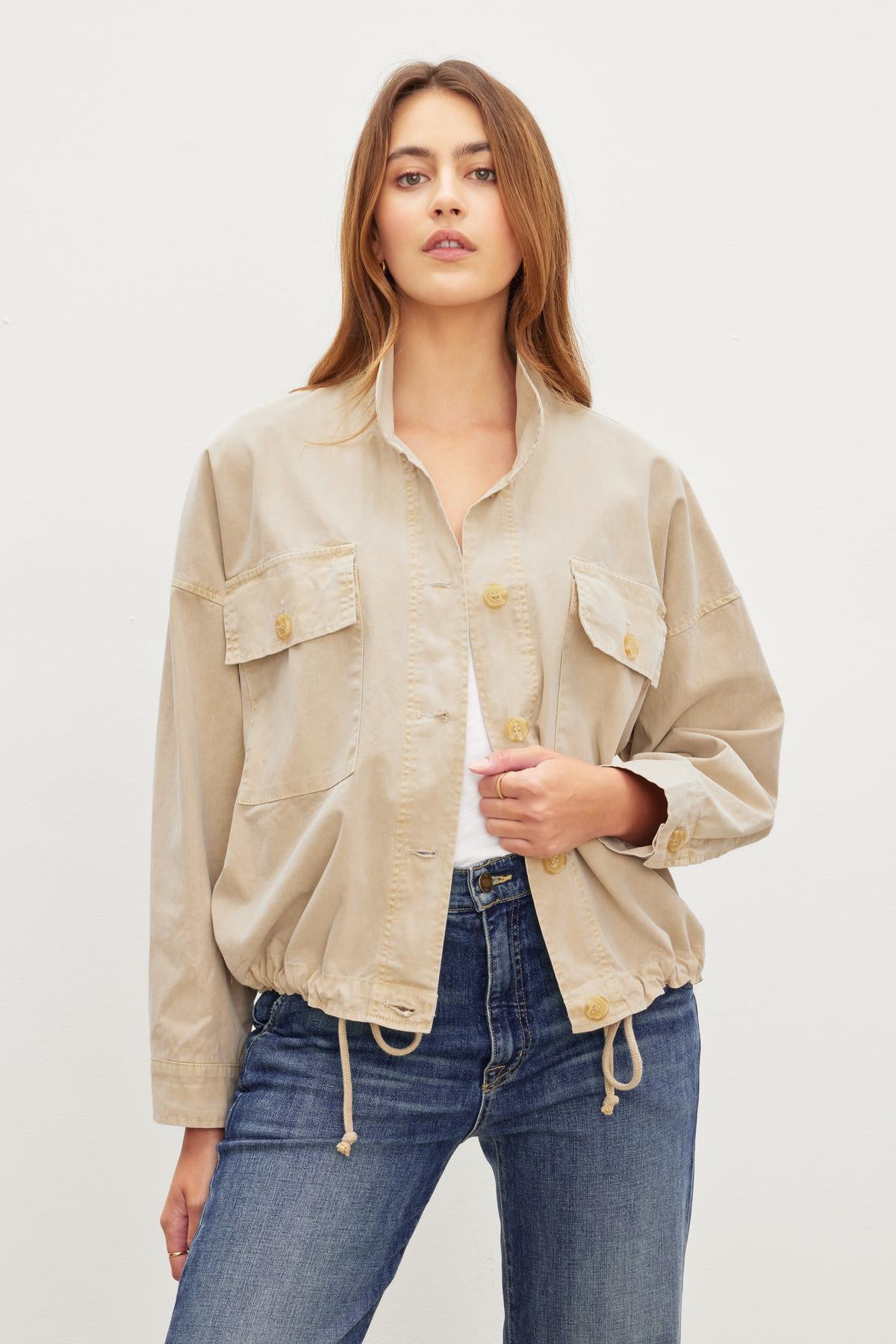A person with long hair wearing a beige, lightweight NICOLA JACKET by Velvet by Graham & Spencer and blue jeans stands against a plain white background.-37229021987009