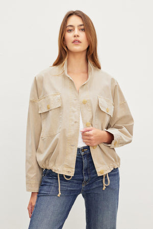 A person with long hair wearing a beige, lightweight NICOLA JACKET by Velvet by Graham & Spencer and blue jeans stands against a plain white background.