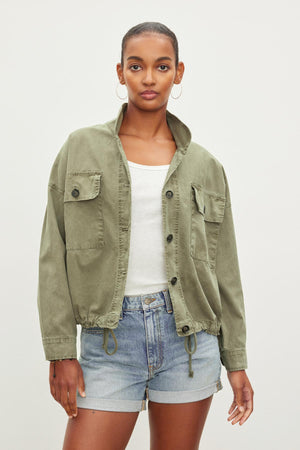 A person wearing an olive green NICOLA JACKET by Velvet by Graham & Spencer over a white tank top and denim shorts stands against a plain background.
