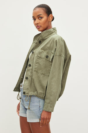 A person wearing a green NICOLA JACKET by Velvet by Graham & Spencer with a cropped hemline and denim shorts stands against a plain background, looking at the camera.