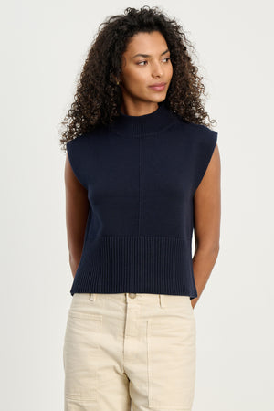 A woman with curly hair stands against a plain background, wearing the Velvet by Graham & Spencer ELECTRA SWEATER VEST—a sleeveless navy blue cotton cashmere blend knit top—paired with light beige pants.