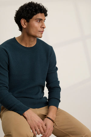 A man with short dark curly hair sits sideways, wearing the FENN SWEATER by Velvet by Graham & Spencer—a webbed texture dark green cotton cashmere crewneck—paired with tan pants, looking off to the side against a plain light background.