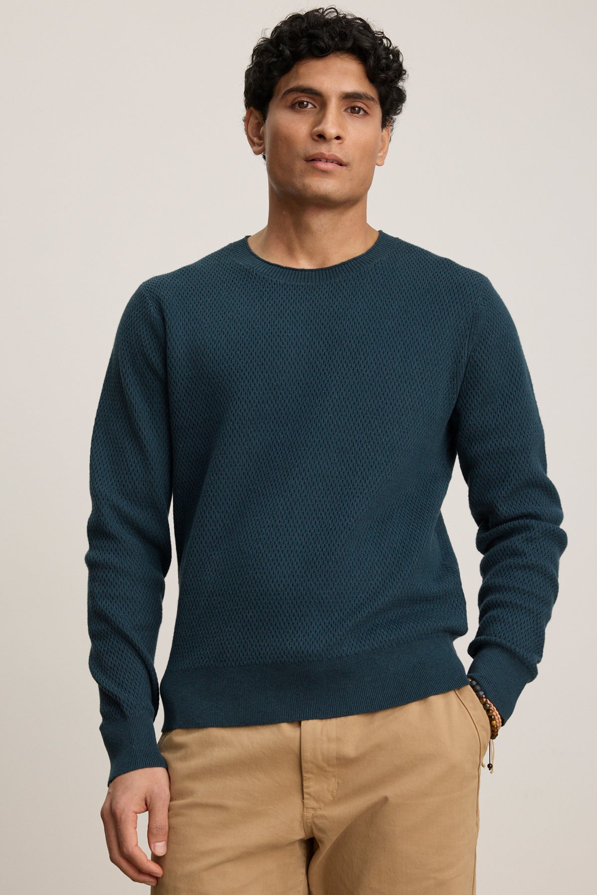 A person stands facing forward, wearing the Velvet by Graham & Spencer FENN SWEATER in teal and tan pants, with a neutral expression and one hand in their pocket against a plain background.