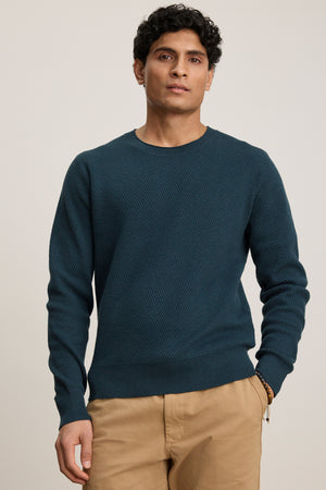 A person stands facing forward, wearing the Velvet by Graham & Spencer FENN SWEATER in teal and tan pants, with a neutral expression and one hand in their pocket against a plain background.