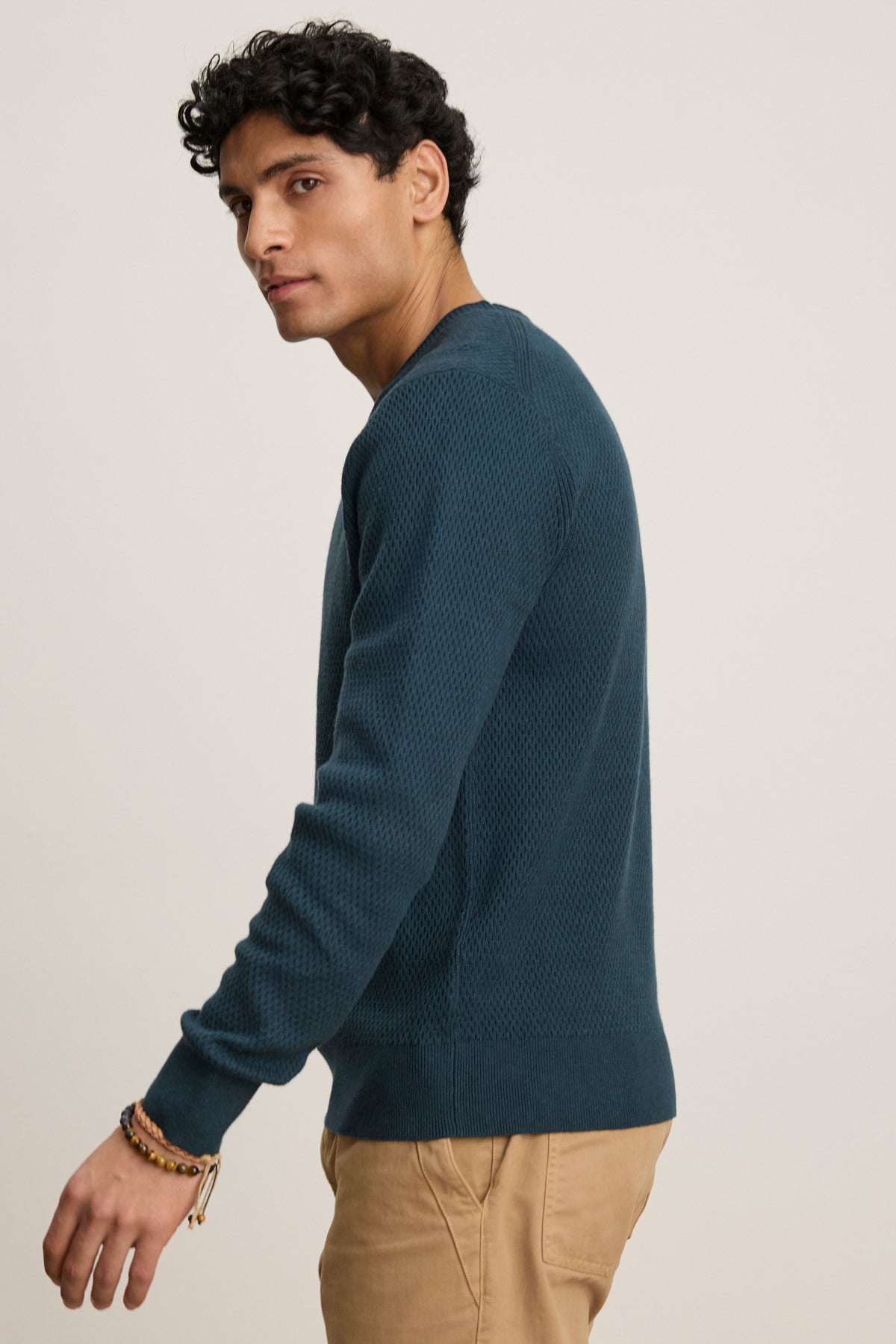A person wearing the FENN SWEATER by Velvet by Graham & Spencer—a blue crewneck with a webbed texture—and beige pants stands against a plain background, facing slightly to the side.