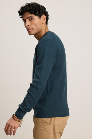 A person wearing the FENN SWEATER by Velvet by Graham & Spencer—a blue crewneck with a webbed texture—and beige pants stands against a plain background, facing slightly to the side.