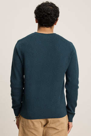 A person faces away from the camera, wearing the FENN SWEATER by Velvet by Graham & Spencer—a webbed texture dark blue crewneck—paired with beige pants against a plain background.
