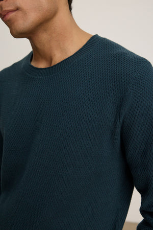 A person wearing the FENN SWEATER by Velvet by Graham & Spencer—a dark teal crewneck with a webbed texture—shown from shoulders to chest.