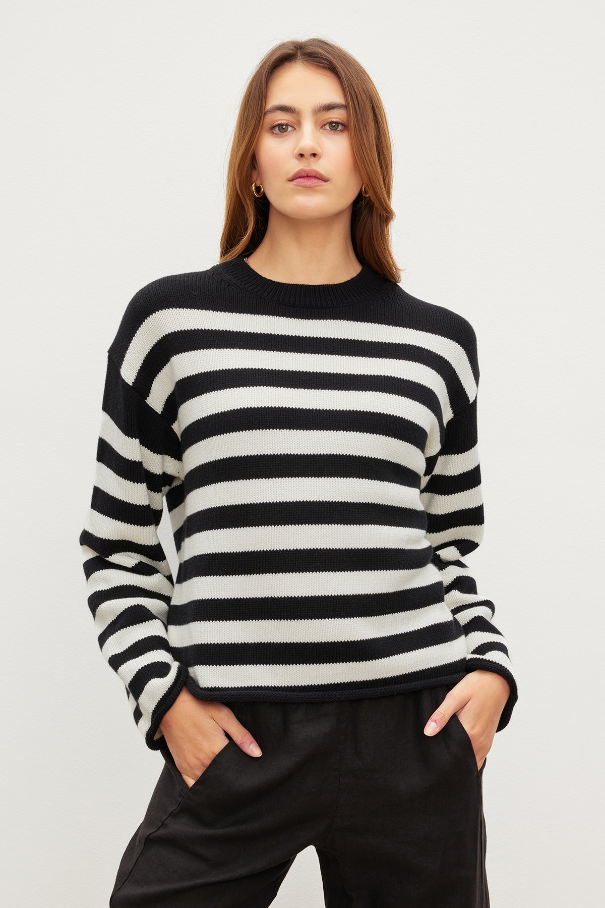 A person stands against a plain background in a LEX COTTON CASHMERE SWEATER by Velvet by Graham & Spencer, featuring a black and white striped design with hands in pockets, exuding an effortless classic Breton charm.-35967621923009