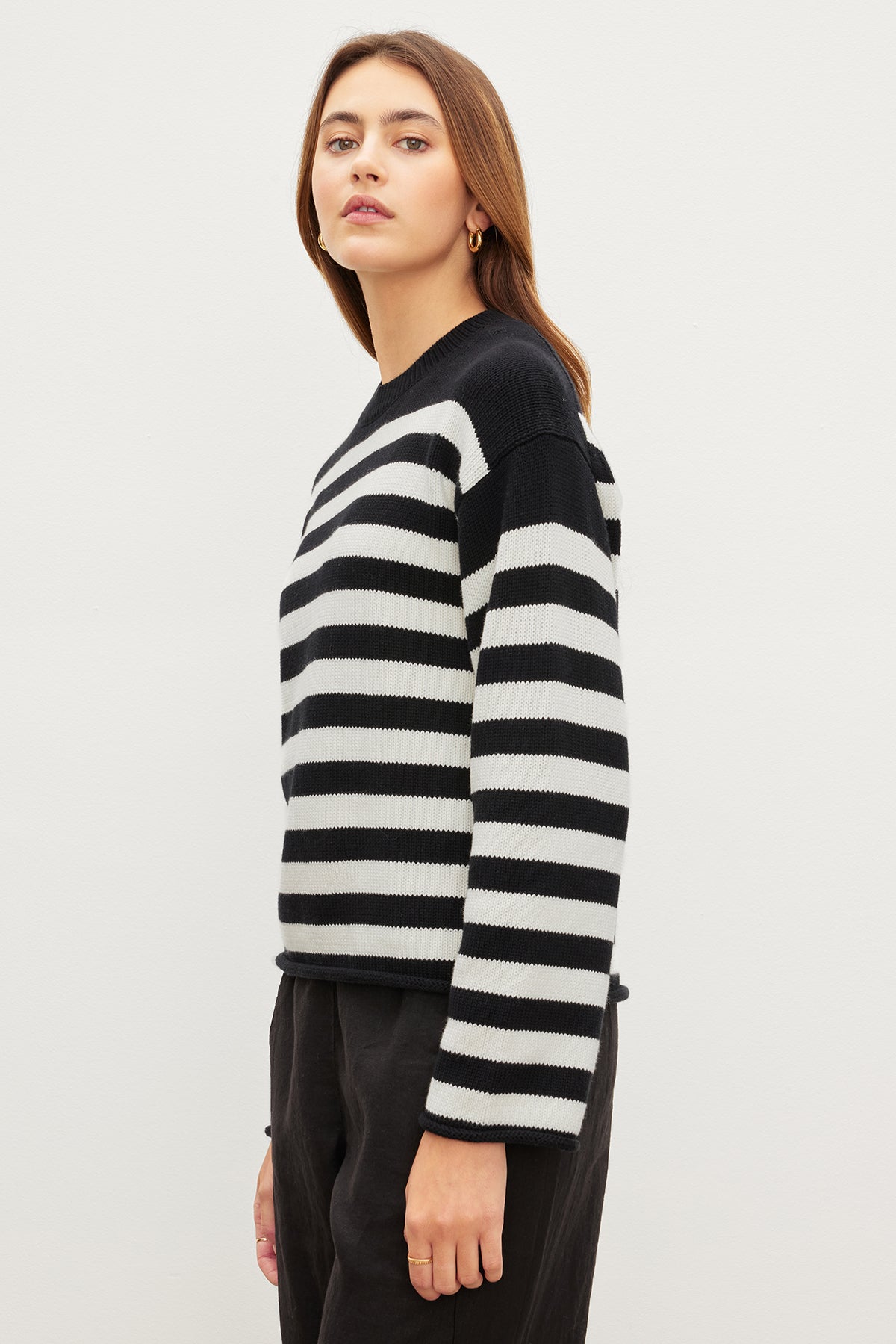 A person exudes a casual vibe in the LEX COTTON CASHMERE SWEATER by Velvet by Graham & Spencer, with its black and white breton stripes, paired with dark pants against a light backdrop, gazing ahead.-35967621857473