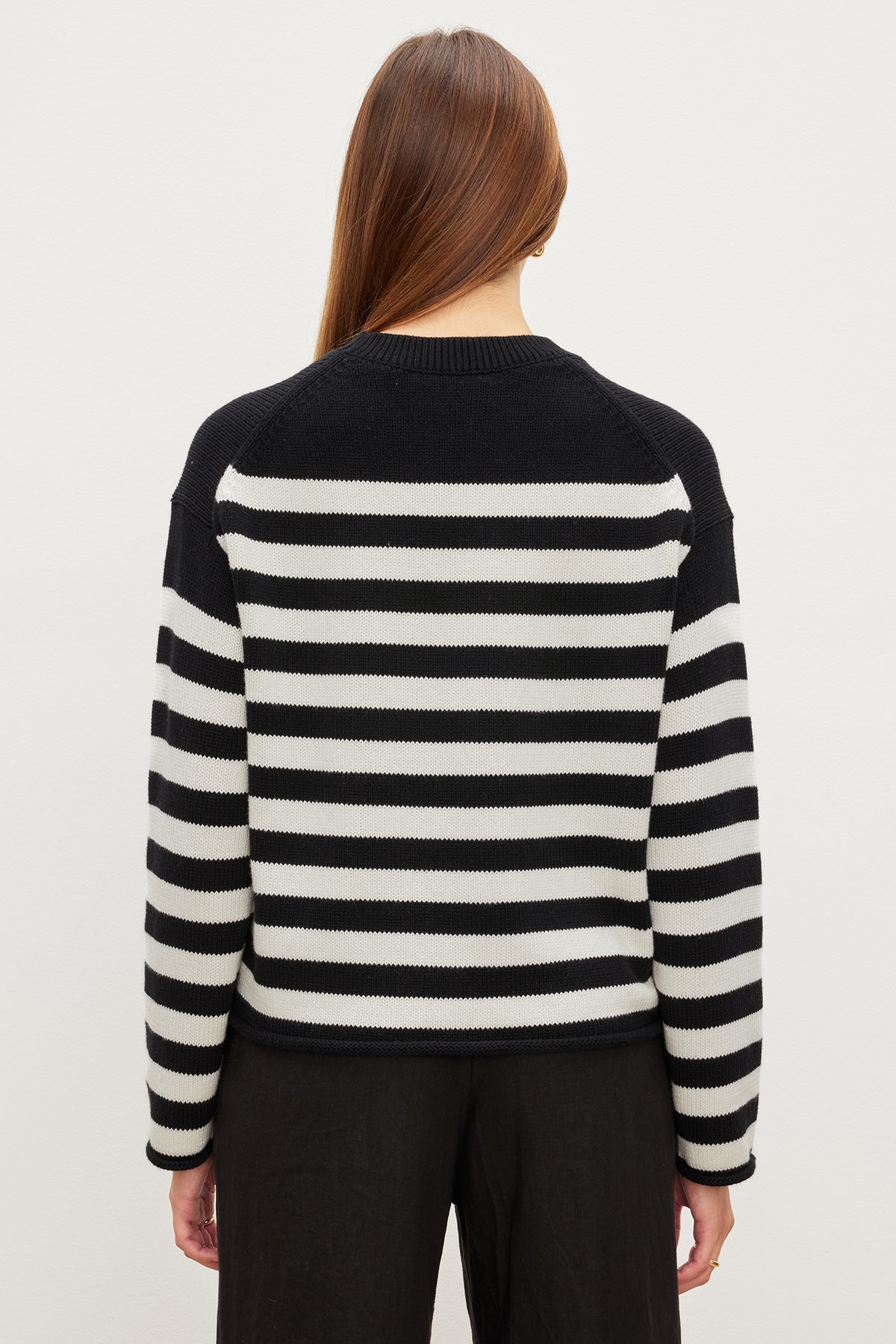 A person with long hair is seen from the back wearing the Velvet by Graham & Spencer LEX COTTON CASHMERE SWEATER, featuring black and white stripes, paired with black pants for a laid-back vibe against a plain background.