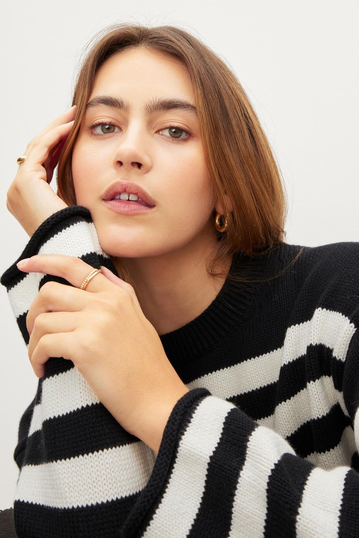 A long-haired person, clad in a black and white LEX COTTON CASHMERE SWEATER by Velvet by Graham & Spencer, reminiscent of a relaxed Breton style, rests their hand on their face while looking forward.-36040376516801