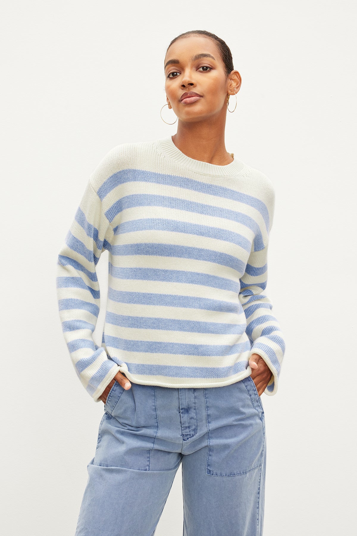 Dressed in a Velvet by Graham & Spencer LEX COTTON CASHMERE SWEATER, this person pairs the laid-back light blue and white stripes with light blue jeans and poses against a plain white backdrop, showcasing the classic Breton silhouette.