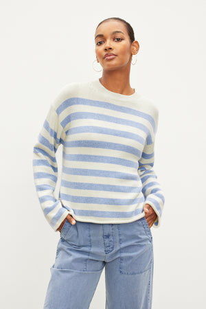 Dressed in a Velvet by Graham & Spencer LEX COTTON CASHMERE SWEATER, this person pairs the laid-back light blue and white stripes with light blue jeans and poses against a plain white backdrop, showcasing the classic Breton silhouette.