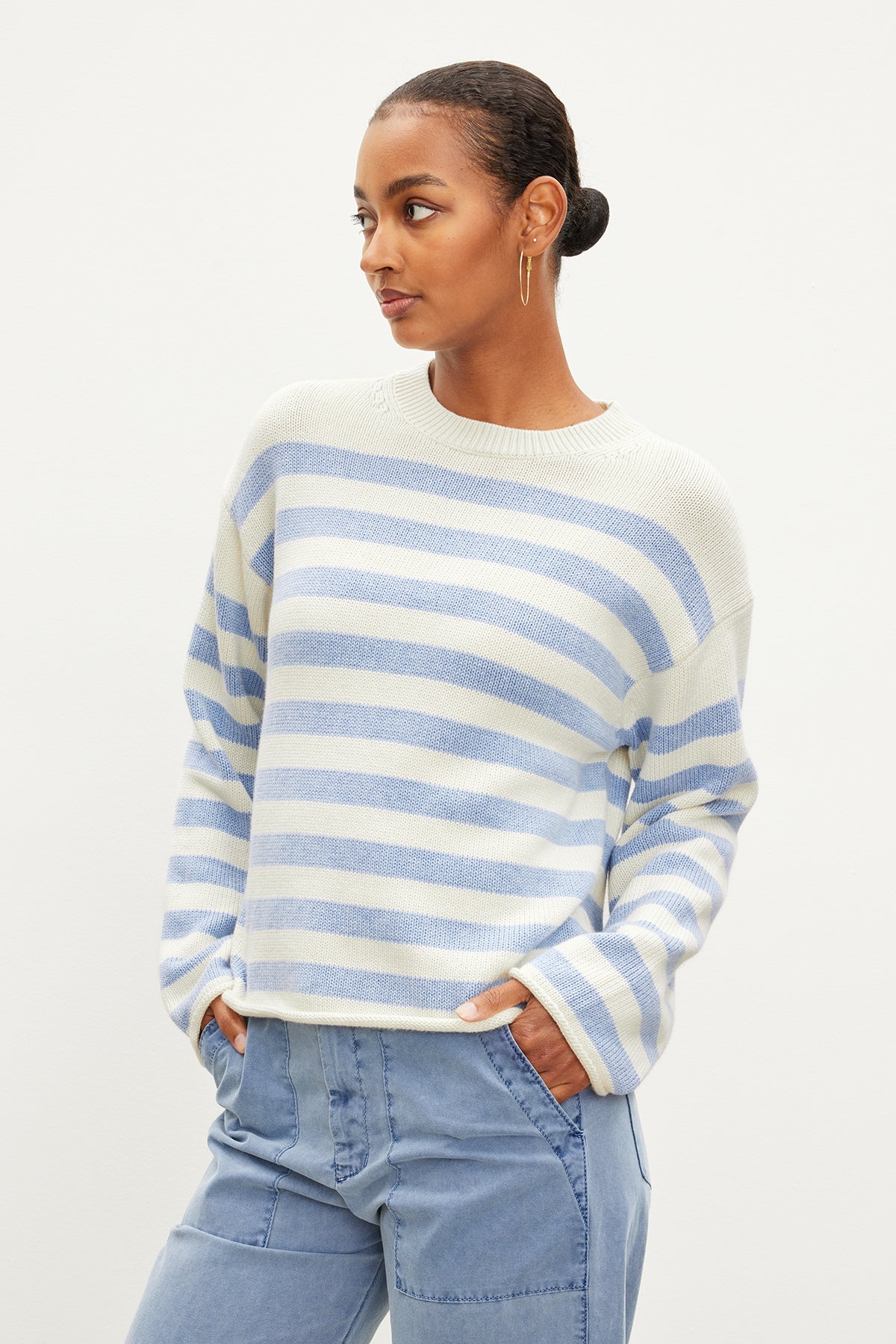 Sporting Velvet by Graham & Spencer's LEX COTTON CASHMERE SWEATER, a light blue and white striped Breton-inspired piece, paired with light blue pants, they embody relaxed elegance. Hands in pockets and short hair styled, they gaze to the side meditatively.-35967621726401