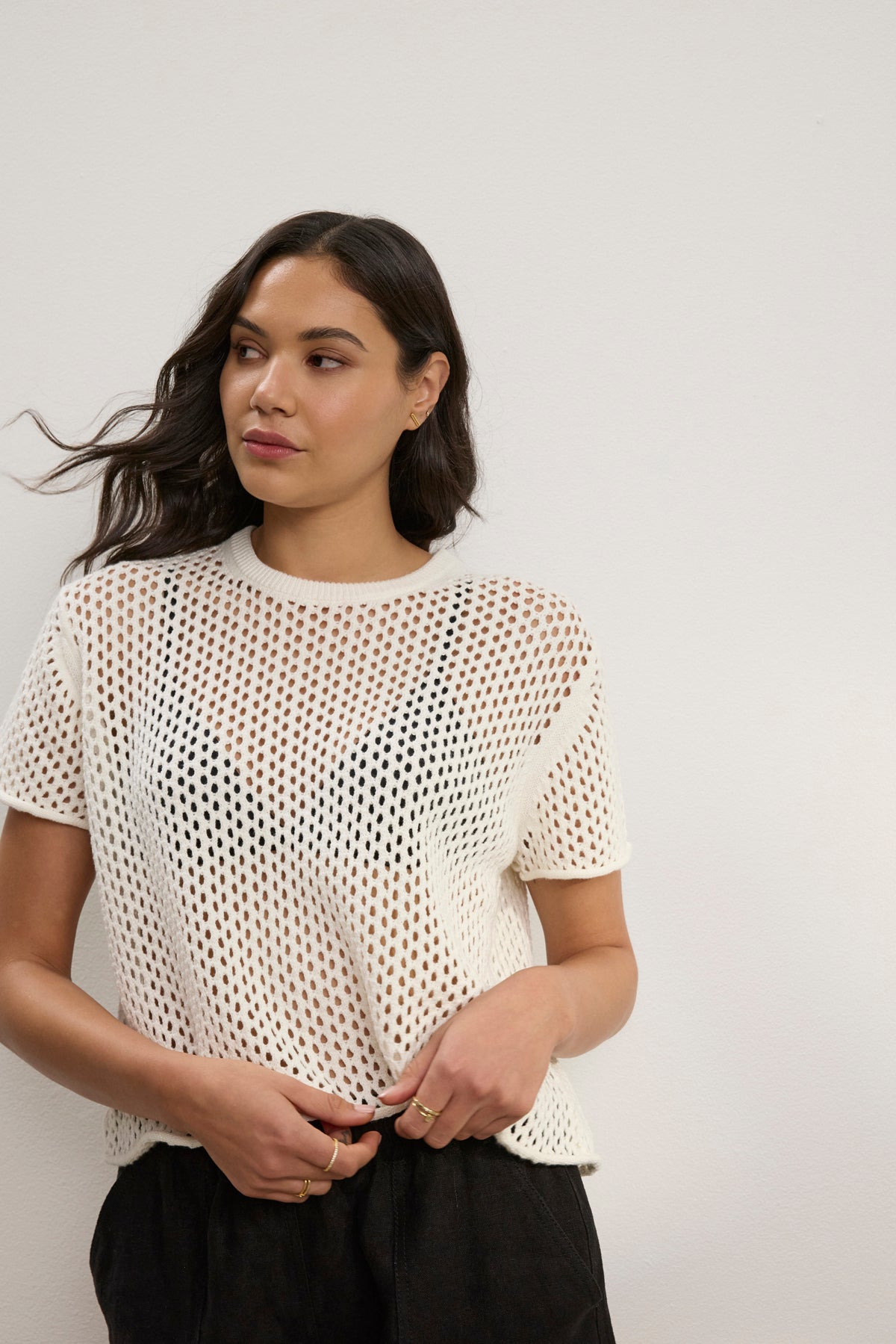 A woman stands against a light wall, wearing Velvet by Graham & Spencer’s ASHLEY COTTON CASHMERE MESH SWEATER in white over an underlayer and black pants, looking to the side with one hand on her waist.-39123268305089