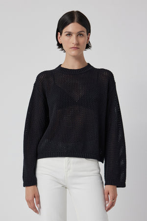 A person with short dark hair is wearing the Velvet by Jenny Graham's KANAN cotton cashmere mesh sweater and white pants, standing against a plain background.
