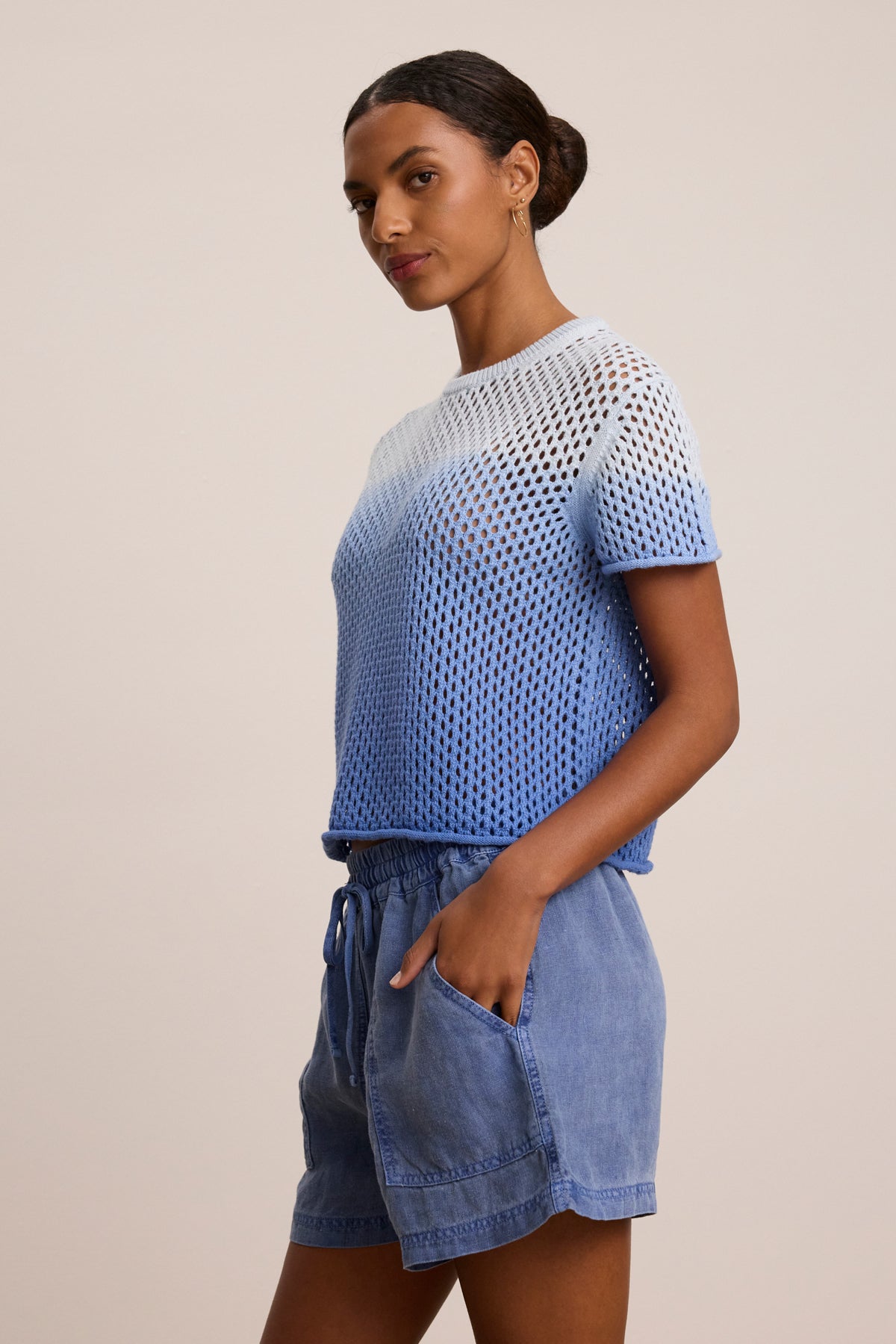 A woman stands against a plain background, wearing the POPPY COTTON CASHMERE MESH SWEATER by Velvet by Graham & Spencer in blue and white, paired with blue drawstring shorts, one hand casually in her pocket.