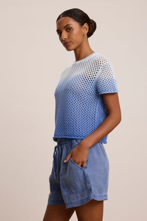 A woman stands against a plain background, wearing the POPPY COTTON CASHMERE MESH SWEATER by Velvet by Graham & Spencer in blue and white, paired with blue drawstring shorts, one hand casually in her pocket.