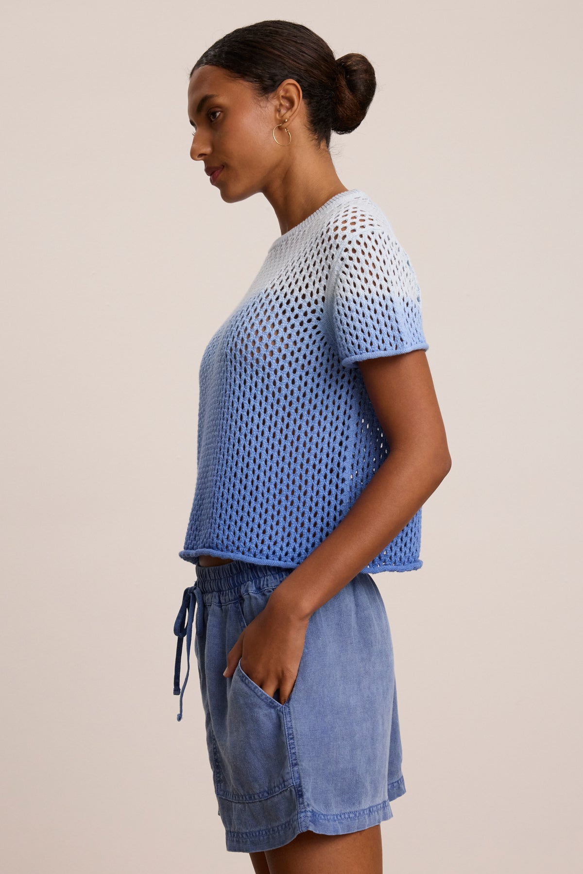 A person in profile wears the POPPY COTTON CASHMERE MESH SWEATER by Velvet by Graham & Spencer, featuring blue and white dip dye short sleeves, paired with light blue drawstring shorts, hands in pockets, against a plain background.
